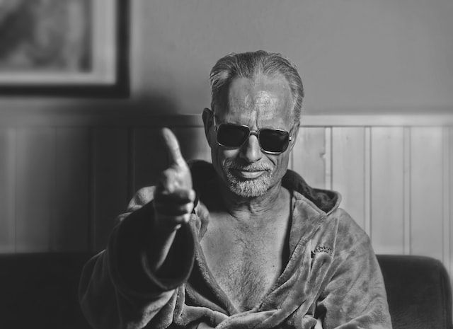 Black and white picture of man wearing a bathrobe open to expose his chest, sunglasses, and a wry grin, holding his hand out to shake hands