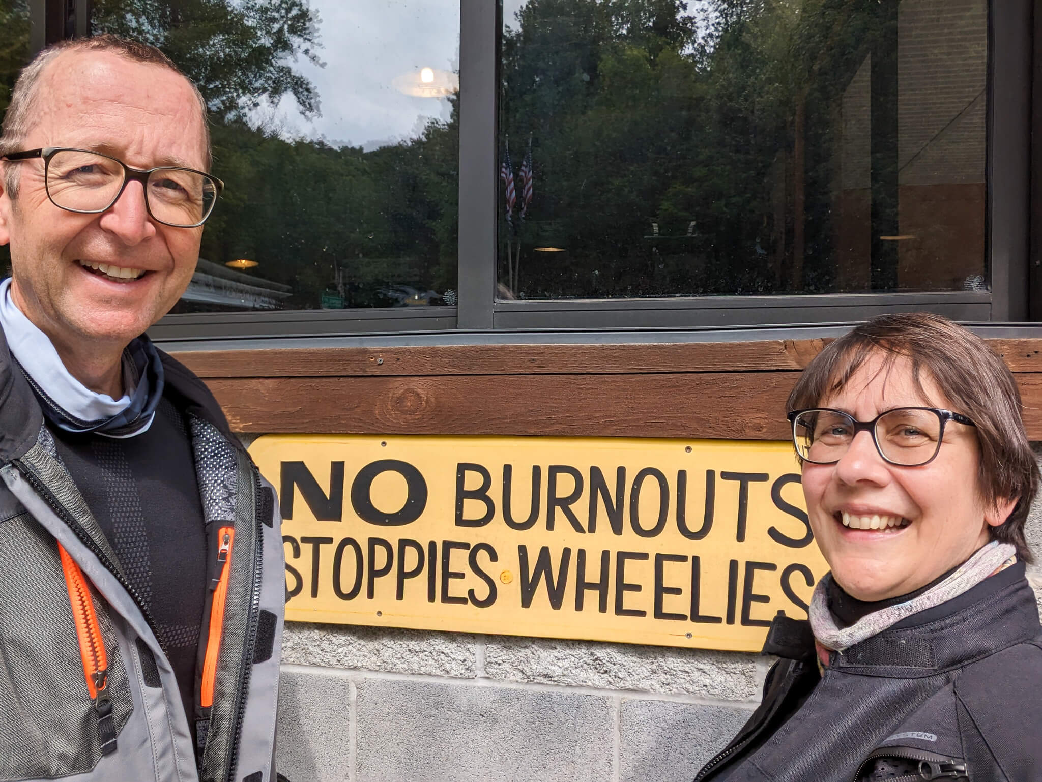 Two riders in front of sign reading “No burnouts, stoppies, wheelies”