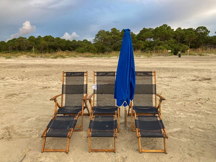 Three lounge chairs on a sandy beach with a blue umbrella (closed) and pine trees in background