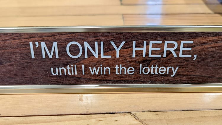 Desk placard reading "I'M ONLY HERE, until I win the lottery". 